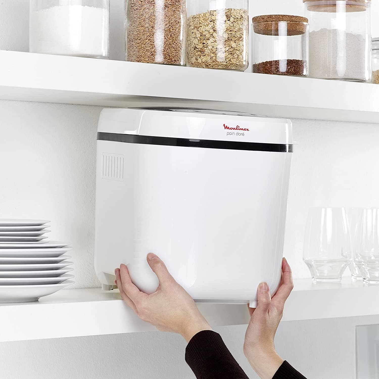 Deux mains rangent la machine à pain Moulinex Pain Doré OW210130 blanche sur une étagère de cuisine, sous des bocaux de farine et de céréales