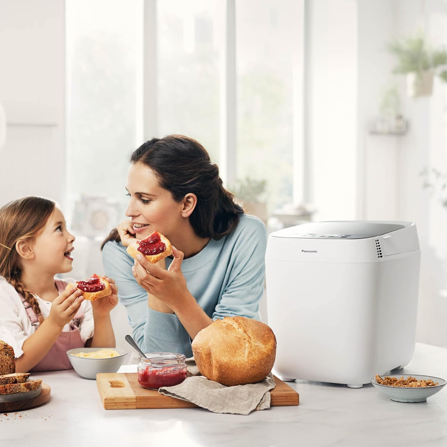 Mère et fille dégustant des tartines de confiture dans une cuisine lumineuse, à côté d'une machine à pain blanche et d'une miche dorée