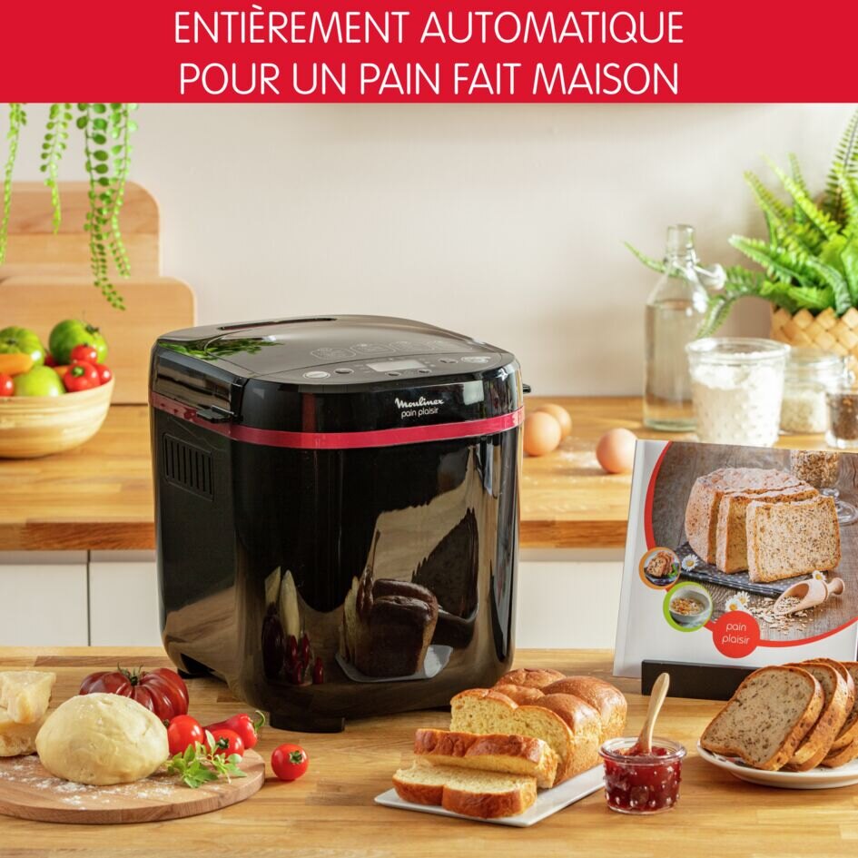 Moulinex Pain Plaisir OW220830 posée sur un plan de travail en bois, entourée d'un pâton, de tranches de brioche, d'un pot de confiture et du livret de recettes